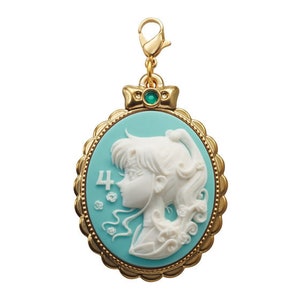 May include: A gold-colored charm with a blue cameo featuring a white silhouette of a woman with long hair. The cameo is surrounded by a gold-colored frame with a decorative edge. The charm has a green gem at the top.