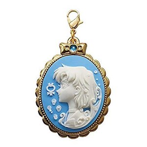 May include: A gold-colored charm with a blue cameo featuring a white silhouette of a woman with long hair. The cameo is surrounded by a gold-colored frame with a decorative border. The charm has a blue gemstone at the top.