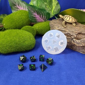 May include: A set of seven black metal dice with green numbers. The dice are in a variety of shapes, including a d4, d6, d8, d10, d12, and d20. The dice are on a blue surface. There is a clear plastic mold for making dice in the background.