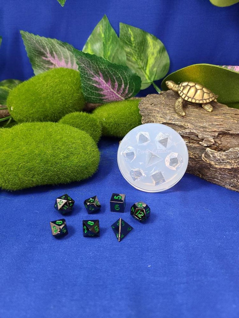 May include: A set of seven black metal dice with green numbers. The dice are in a variety of shapes, including a d4, d6, d8, d10, d12, and d20. The dice are on a blue surface. There is a clear plastic mold for making dice in the background.