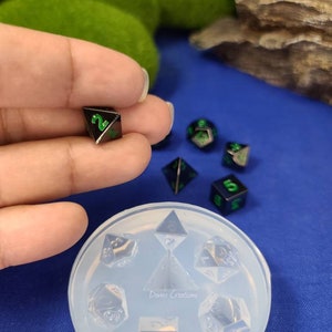 May include: A clear silicone mold for making dice. The mold has seven impressions, one for each face of a standard die. The mold is sitting on a blue surface. There are six black and green dice on the surface next to the mold.