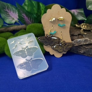 May include: A clear silicone mould with two butterfly shapes and a few smaller shapes.  The mould is next to a wooden display card with two pairs of butterfly earrings.  The earrings are gold with black and glitter accents.  The text "Denis Creations" is visible on the mould.