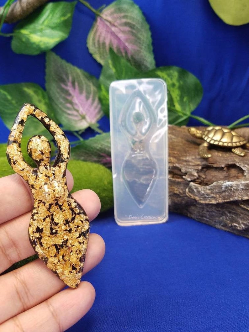 以下が含まれることがあります： 青い背景に、腕を上げた女性の金色のレジン製フィギュア。フィギュアは金箔でできており、手のひらに乗っています。画像には、同じフィギュアの透明なシリコンモールドも写っています。