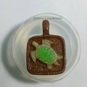 Pode incluir: Um pingente de cor cobre com um design de tartaruga verde e branca. O pingente está em um molde de plástico transparente.
