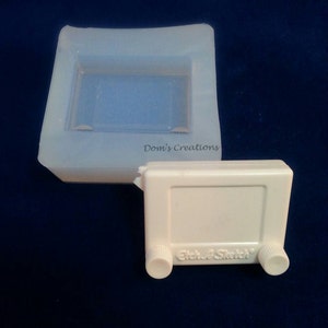 May include: A white plastic Etch A Sketch toy with two knobs on the side. The toy is in a clear silicone mold.