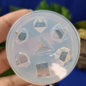 May include: A clear silicone mold for making resin dice. The mold has six cavities, each shaped like a different type of die. The mold is sitting on a hand.