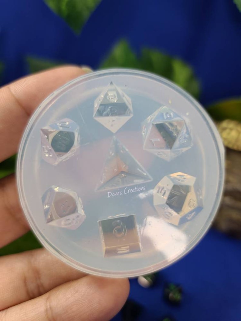 May include: A clear silicone mold for making resin dice. The mold has six cavities, each shaped like a different type of die. The mold is sitting on a hand.