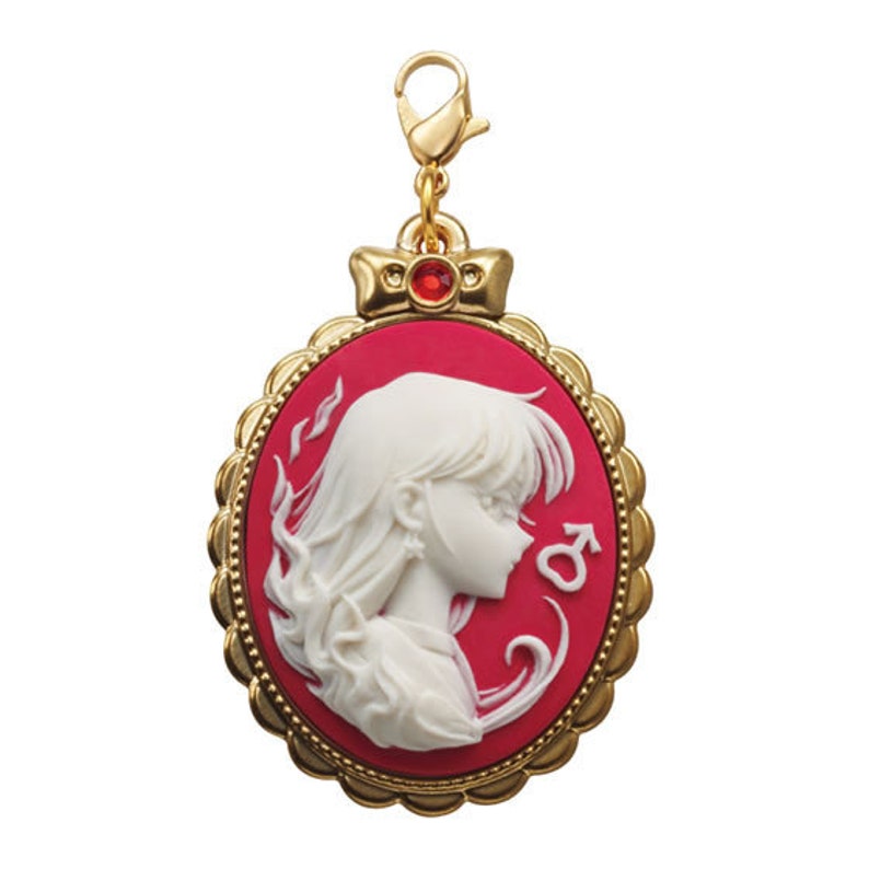 May include: A gold-colored charm with a red cameo featuring a white portrait of a woman with long hair. The cameo is surrounded by a gold-colored frame with a decorative edge. The charm has a red gemstone at the top.