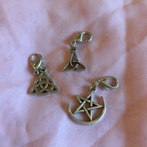May include: Three silver charm pendants with a silver clip. The charms are a triquetra, a pentagram with a crescent moon, and another triquetra.