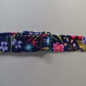 K&ouml;nnte beinhalten: Ein marineblauer Stoffkragen mit einem Blumenmuster mit rosa, lila und gelben Blumen. Der Kragen hat einen Metall-Schnallenverschluss. Das florale Design ist vor einem dunkelblauen Hintergrund angeordnet.
