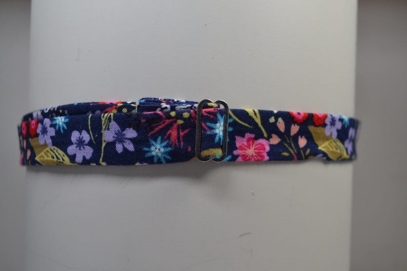 K&ouml;nnte beinhalten: Ein marineblauer Stoffkragen mit einem Blumenmuster mit rosa, lila und gelben Blumen. Der Kragen hat einen Metall-Schnallenverschluss. Das florale Design ist vor einem dunkelblauen Hintergrund angeordnet.