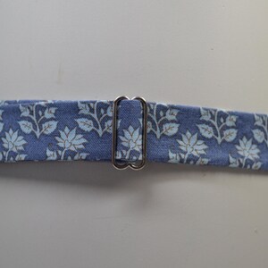 K&ouml;nnte beinhalten: Ein blauer Stoffg&uuml;rtel mit einem floralen Muster in Hellblau und Wei&szlig;. Der G&uuml;rtel hat eine silberfarbene Metallschnalle. Der G&uuml;rtel ist um einen wei&szlig;en zylindrischen Gegenstand gewickelt.