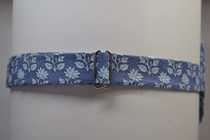 K&ouml;nnte beinhalten: Ein blauer Stoffg&uuml;rtel mit einem floralen Muster in Hellblau und Wei&szlig;. Der G&uuml;rtel hat eine silberfarbene Metallschnalle. Der G&uuml;rtel ist um einen wei&szlig;en zylindrischen Gegenstand gewickelt.