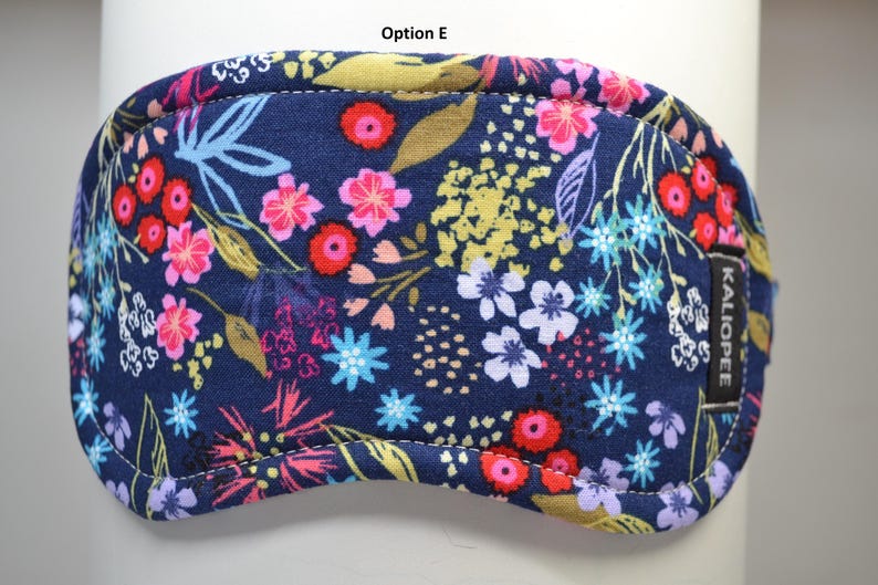K&ouml;nnte beinhalten: Eine marineblaue Schlafmaske mit einem farbenfrohen Blumenmuster. Das Design zeigt rosa, rote, gelbe und blaue Blumen und Bl&auml;tter. Die Maske hat einen gen&auml;hten Rand und ein kleines schwarzes Etikett mit der Aufschrift "KALIOPEE".