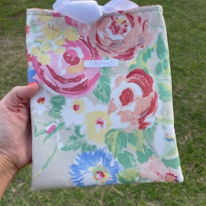 May include: A floral print case with a white bow. The case features a pattern of large pink, red, yellow, and blue flowers on a beige background. The text "Quilted Morning Co." is visible on a small white label.