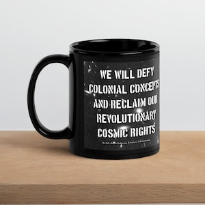 Könnte beinhalten: Eine schwarze Keramik-Tasse mit weißem Text. Der Text auf der Tasse lautet "WE WILL DEFY COLONIAL CONCEPTS AND RECLAIM OUR REVOLUTIONARY COSMIC RIGHTS." Die Tasse steht auf einer hellbraunen Holzoberfläche.