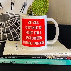 Könnte beinhalten: Eine weiße Kaffeetasse mit einem roten Band um den oberen Rand. Die Tasse trägt den Text "We will continue to fight for a decolonized future paradise". Die Tasse steht auf einem Stapel Bücher mit den Titeln "Wet Land", "Lucas de Lima", "Indecency", "Coffee House Press" und "Action Books".