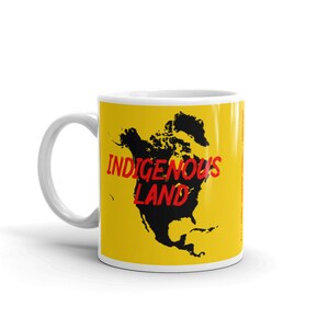 Könnte beinhalten: Eine weiße Keramiktasse mit gelbem Hintergrund und einer schwarzen Silhouette von Nordamerika. Der Text "INDIGENOUS LAND" ist in Rot auf die Tasse gedruckt.