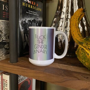 Könnte beinhalten: Weiße Keramik-Tasse mit dem Text "FUCK THE GENDER BINARY" in Schwarz. Die Tasse hat einen Hintergrund mit Pastellfarben. Die Tasse steht auf einem Holzregal mit Büchern und Dekorationsartikeln.