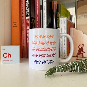 Könnte beinhalten: Eine weiße Keramiktasse mit einem Regenbogen-Farbverlauf. Der Text auf der Tasse lautet "In a dream you saw a way to decolonize and you were full of joy."