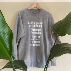 May include: Gray sweatshirt with white text that reads "Love & protect yr community Dismantle the empire Honor yr ancestors & future generations"