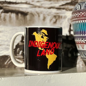Könnte beinhalten: Eine schwarze Kaffeetasse mit einer gelben Umrandung der Weltkarte und dem Text "Indigenous Land" in roten Buchstaben.