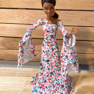 Puede incluir: Muñeca de moda con un vestido largo con estampado floral y mangas acampanadas. El vestido presenta un escote cuadrado y un dobladillo con volantes. La tela es blanca con un estampado de flores rosas, naranjas y moradas.