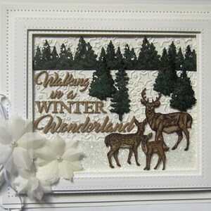 May include: A white card with a white frame and a white border. Inside the frame is a glittery white background with a brown deer family, green trees, and the text "Walking in a Winter Wonderland".