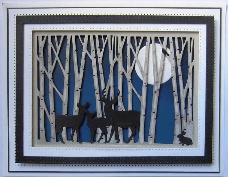 May include: A paper craft scene depicting a moonlit forest with a full moon, a flock of birds flying overhead, and a family of deer standing in the foreground. The forest is made of white paper with a blue background.