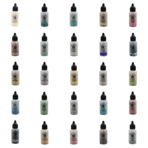 Peut inclure: Vingt-cinq flacons de colle à paillettes de différentes couleurs, dont l'or, l'argent, le vert, le bleu, le violet et le rose. Chaque flacon a un bouchon noir et une étiquette blanche avec les mots "Magic Glitter Glue" et un cercle avec un motif en spirale.
