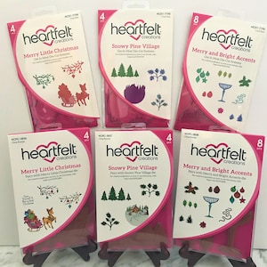 May include: Six packages of Heartfelt Creations Christmas themed die-cutting stamps. The packages are pink and feature a variety of Christmas designs, including trees, ornaments, reindeer, and a sleigh. The packages are labeled with the names of the dies, such as "Merry Little Christmas" and "Snowy Pine Village".