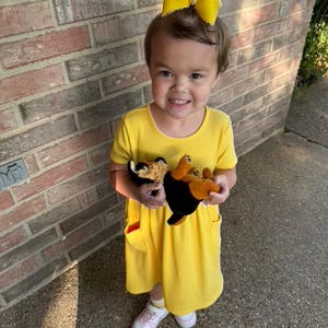Pu&ograve; includere: Un bambino piccolo indossa un vestito giallo brillante con tasche e un fiocco giallo abbinato tra i capelli. Il bambino tiene in mano due peluche, uno nero e marrone e l'altro marrone e beige. Il bambino indossa scarpe da ginnastica bianche e rosa.