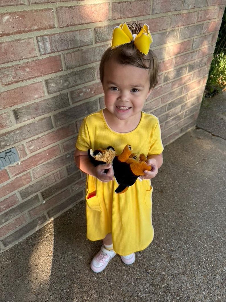 Pu&ograve; includere: Un bambino piccolo indossa un vestito giallo brillante con tasche e un fiocco giallo abbinato tra i capelli. Il bambino tiene in mano due peluche, uno nero e marrone e l'altro marrone e beige. Il bambino indossa scarpe da ginnastica bianche e rosa.