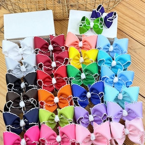 Pu&ograve; includere: Una collezione di fiocchi per capelli colorati in varie tonalit&agrave;, tra cui rosa, blu, verde e viola. Ogni fiocco presenta un bordo bianco e un nodo centrale, disposti in scatole su una superficie di legno. I fiocchi sono di colori solidi e sono ben presentati.