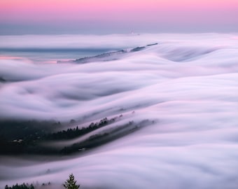 Puede incluir: Una vista panorámica de un paisaje con colinas onduladas cubiertas por una gruesa capa de niebla blanca. La niebla está iluminada por una suave luz rosa del cielo, creando una atmósfera de ensueño y etérea.