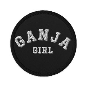 May include: A round, black embroidered patch with the words "GANJA GIRL" in white, arched text. The patch has a black border and is likely designed to be sewn or ironed onto clothing or accessories.