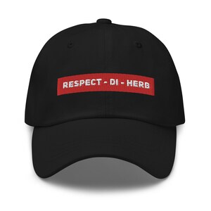 May include: Black baseball cap with a curved brim. The cap features the embroidered text "RESPECT - DI - HERB" in white on a red rectangular patch. The cap is made of a soft fabric and has a classic, casual style.