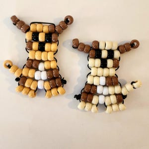 Könnte beinhalten: Zwei handgefertigte Hunde-Figuren aus Perlen. Jede Figur ist aus beigen, braunen, schwarzen und weißen Perlen gefertigt, die auf schwarzem Gummiband aufgefädelt sind. Die Hunde haben ausgestreckte Arme und eingerollte Schwänze und werden auf einer weißen Oberfläche gezeigt.