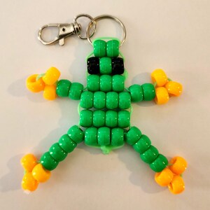 May include: A handmade beaded frog keychain. The frog is made of green beads with black beads for sunglasses and yellow beads for the feet and hands. The keychain has a silver clasp and ring.