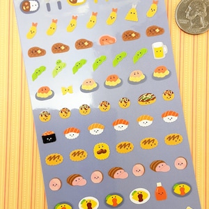 May include: A sheet of colorful stickers featuring various Japanese foods, including sushi, ramen, curry rice, and edamame. The stickers are on a light blue background with the text "Taberaretai" and "Made in Japan".