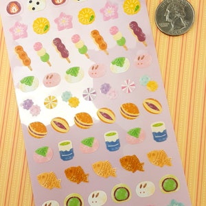 May include: A sheet of stickers featuring various Japanese food and cultural icons. The stickers are on a light purple background and include images of dango, mochi, taiyaki, and flowers. The sheet is labeled "Sticker Selection" in Japanese and English. The sheet also includes the text "Made in Japan".