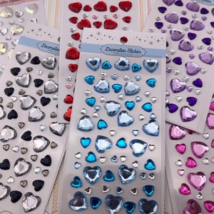 May include: Five sheets of heart-shaped adhesive stickers in various colors including iridescent, black, red, blue, purple, and pink. Each sheet features a header labeled Decoration Sticker with star accents and contains multiple sizes of gems.