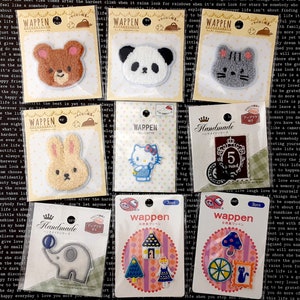 Op de afbeelding: Een assortiment kleurrijke strijkpatches met verschillende cartoonfiguren, dieren en ontwerpen. De patches zijn verpakt in doorzichtige plastic zakjes met witte en gele labels met de tekst "Wappen" en "Handmade".
