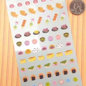 May include: A sheet of Japanese food themed stickers with a variety of colorful designs. The stickers feature cute faces and include items like fish cakes, mochi, dango, and rice balls. The sheet is labeled "Taberaretai" which means "I want to eat it" in Japanese.