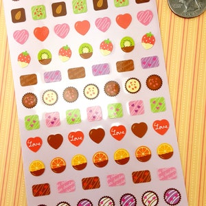 May include: A sheet of pink and white stickers featuring various chocolate-themed designs. The stickers include hearts, squares, circles, and other shapes with different chocolate fillings, such as almonds, strawberries, kiwi, and orange slices. The text "chocotto seal series" and "Love" appears on the sheet.