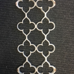 Puede incluir: Una celosía decorativa de metal plateado con un patrón de trébol repetido. La celosía está hecha de metal delgado y tiene un diseño delicado e intrincado.