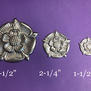 Puede incluir: Tres adornos decorativos plateados en forma de flor de diferentes tamaños. El más grande mide 8,9 cm, el mediano 5,7 cm y el más pequeño 3,8 cm. Se muestran sobre un fondo morado.