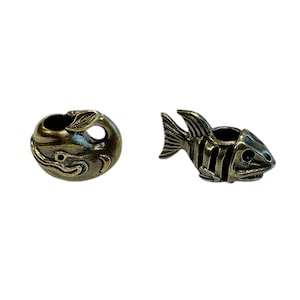 May include: Two metal charms in the shape of fish. One charm is a whale with a hole in the top. The other charm is a fish with a skeleton design and a hole in the top.