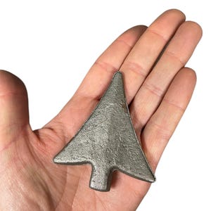 May include: A silver-coloured, arrow-shaped metal object. The arrowhead is triangular, with a small rectangular base. The object is held in a hand against a white background. The metal has a textured surface.