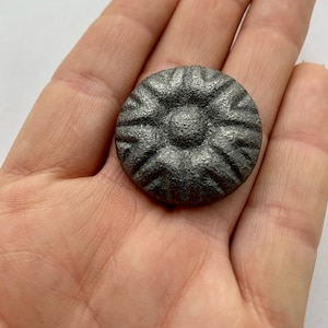 May include: A silver-colored metal button with a raised, starburst design. The button has a textured surface and a slightly rounded shape.